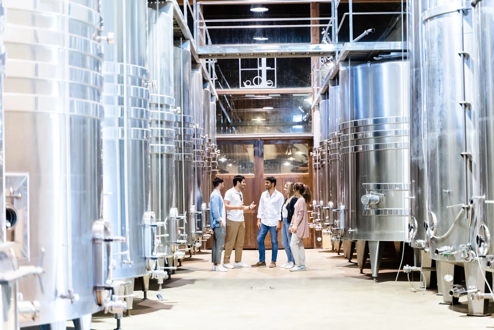 A group of people on a tour, standing between large stainless steel fermentation tanks in a modern winery.