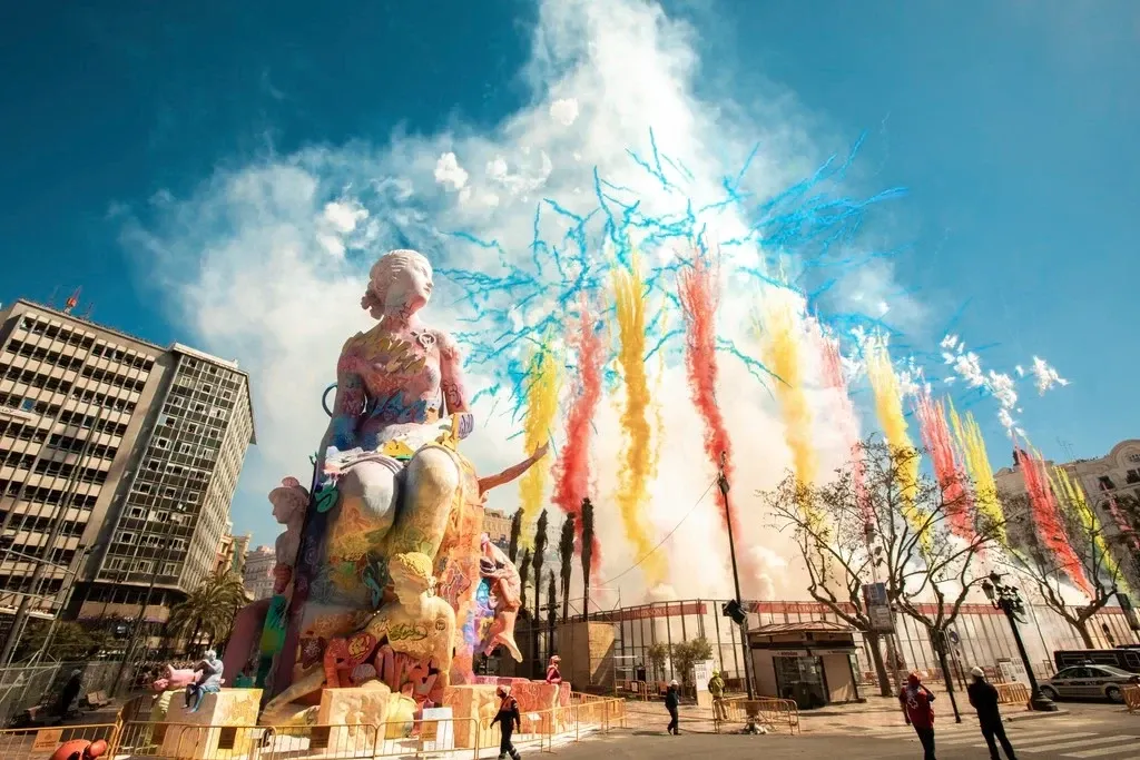 A large, colourful 'falla' statue with a daytime firework display of coloured smoke exploding behind it in a city square.