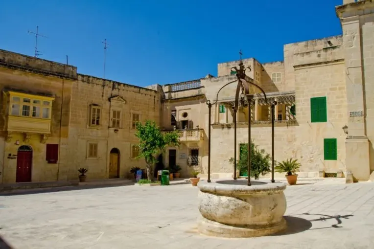 Empty stone square with a historic stone well in the foreground. Traditional buildings with balconies and colorful shutter...