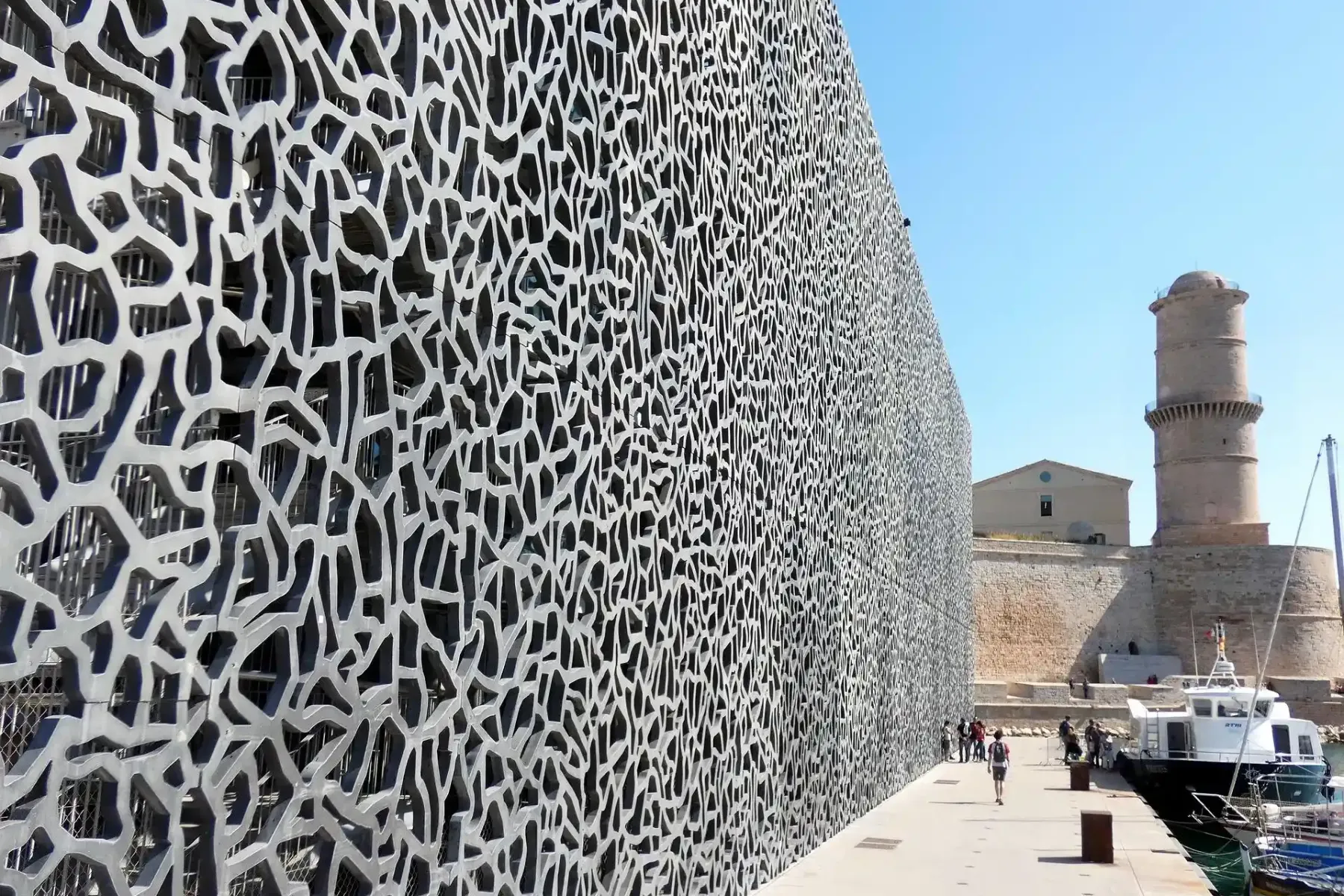 Modern building with intricate, net-like facade and old stone fort under a clear blue sky on a sunny day in Marseille.