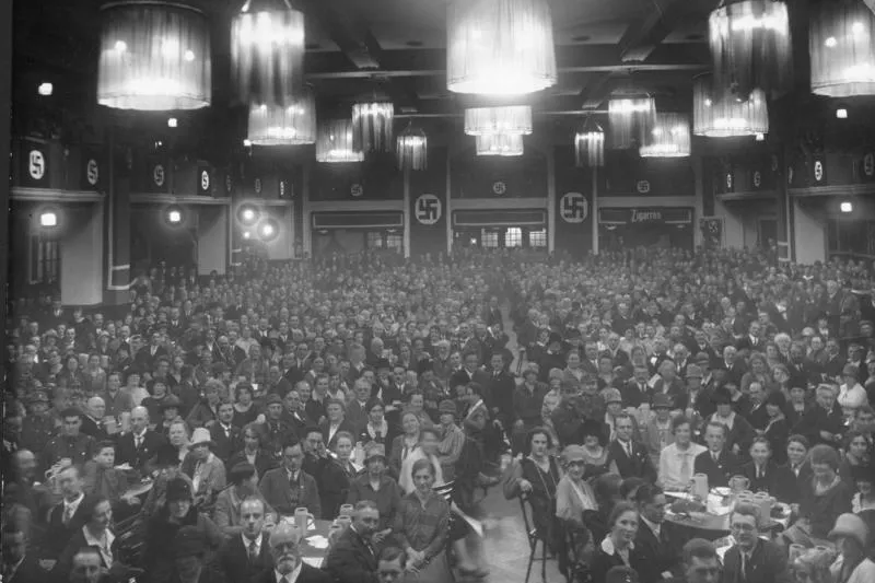 Black and white photo of a large crowded hall filled with people seated at tables. Swastika banners and 'Zigaretten' sign ...