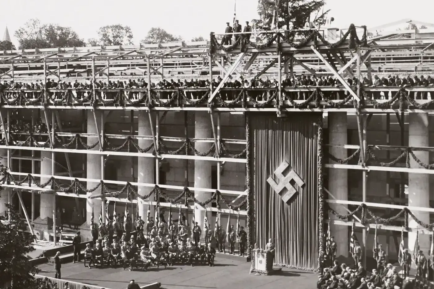 Black and white photo of a building under construction, decorated with garlands and a large swastika banner. People are ga...