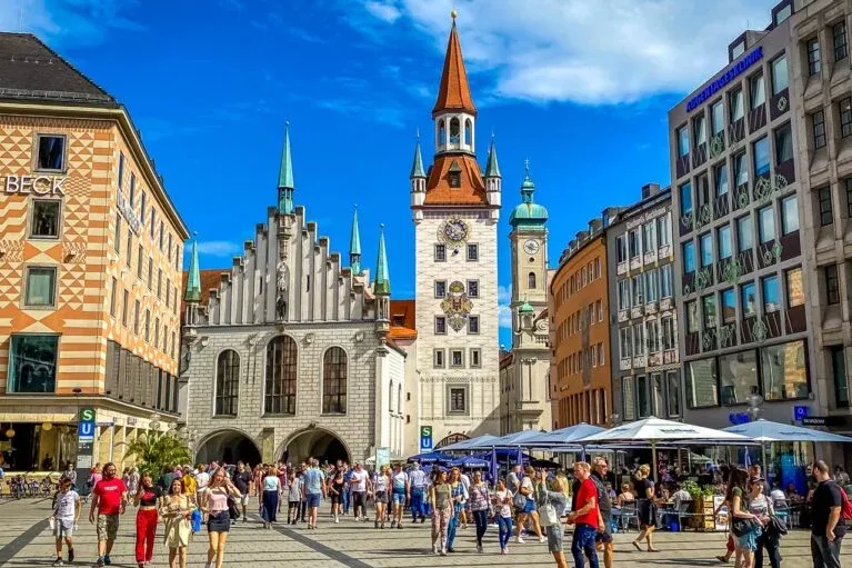 Crowded Marienplatz in Munich with the Old Town Hall and the tower of the New Town Hall under a blue sky.