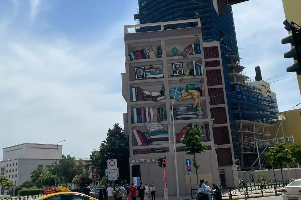 Building with a trompe l'oeil bookshelf mural, a construction skyscraper behind it, a street with a yellow taxi, scooter, ...