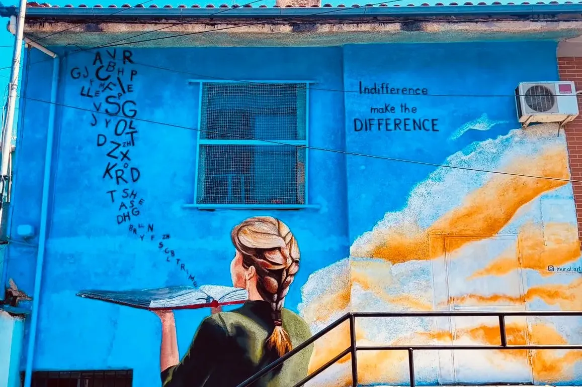Mural on a blue building in Tirana depicting a woman holding an open book with letters flying out, and the text 'Indiffere...