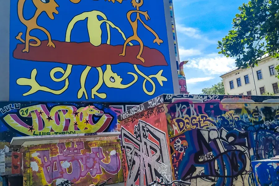 A bright blue building wall with a large yellow and red abstract mural. Below, walls covered in colorful graffiti, parked ...