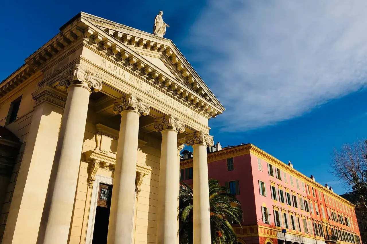 Neo-classical chapel with columns, pediment, and statue under a blue sky, next to a pink building with arcades in Nice.