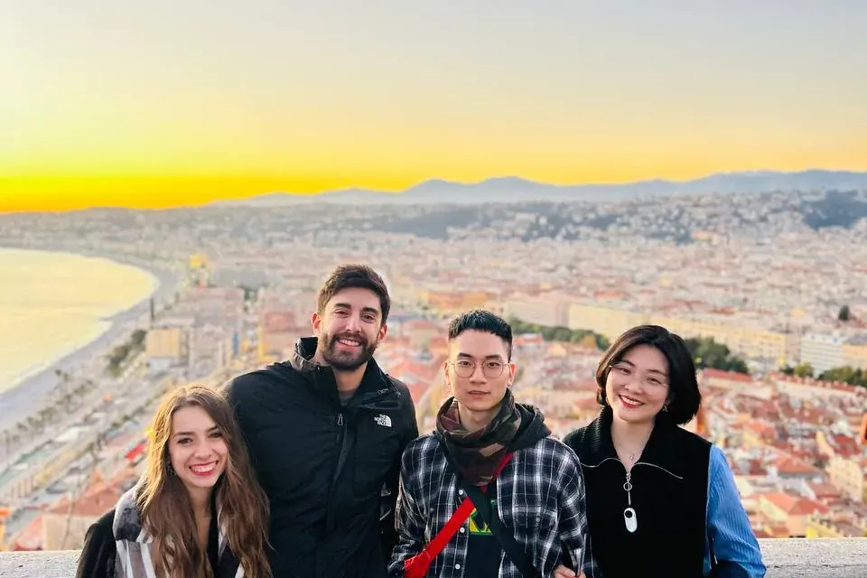 Four people posing on a viewpoint overlooking Nice city, the bay, and mountains at sunset. Golden light illuminates the ho...