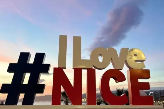 Large '#ILoveNICE' sign illuminated at dusk, with a soft pink and blue sky over the city and sea in Nice.