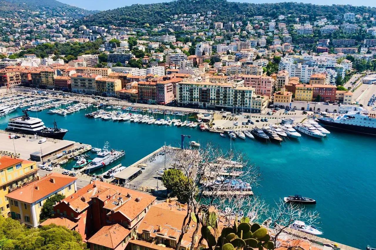 Panoramic view of Nice's Port Lympia with numerous yachts, colorful waterfront buildings, and green hills under a clear bl...