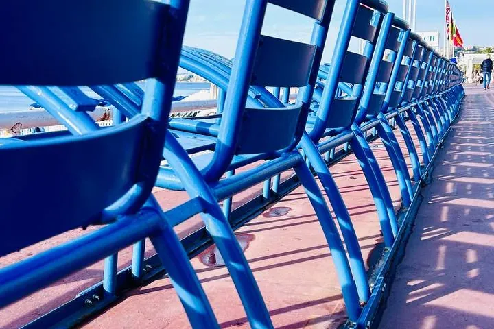 A row of iconic blue chairs on the Promenade des Anglais in Nice, facing the sea under a clear blue sky. Shadows extend on...