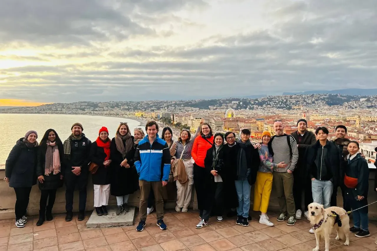 Diverse group and a golden retriever dog on a tiled terrace overlooking Nice city, bay, and coastline at sunset.