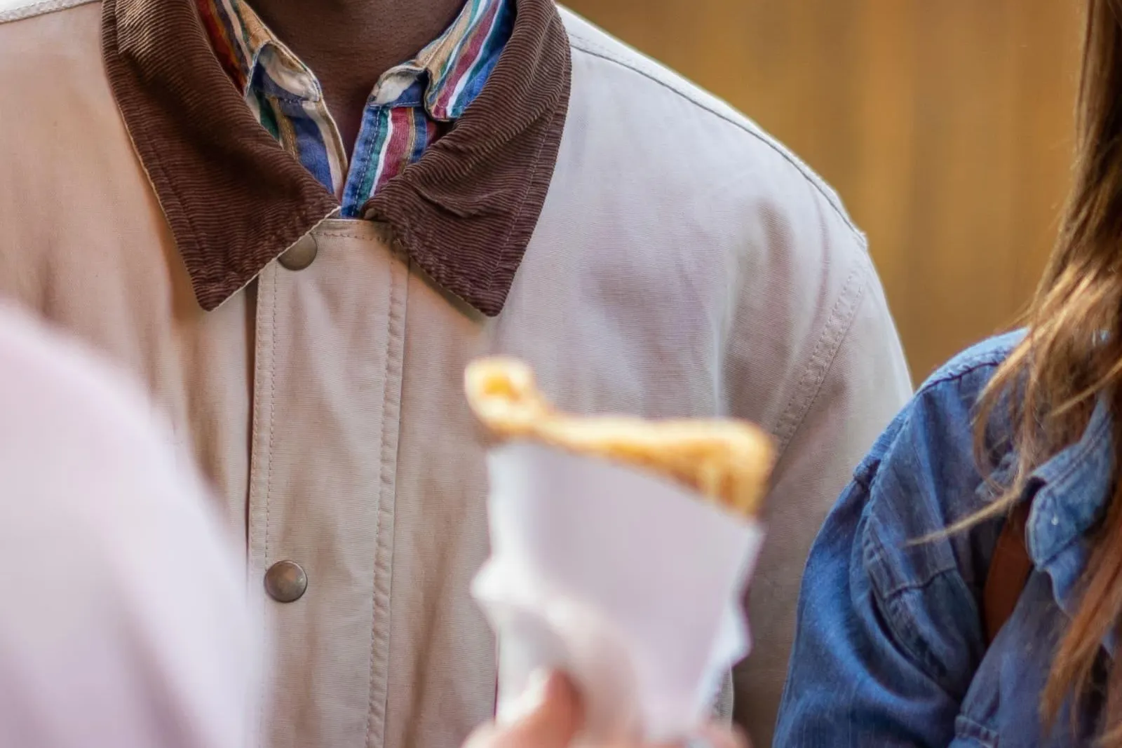 Smiling man watching a crepe being handed to a person in a denim jacket on a street.