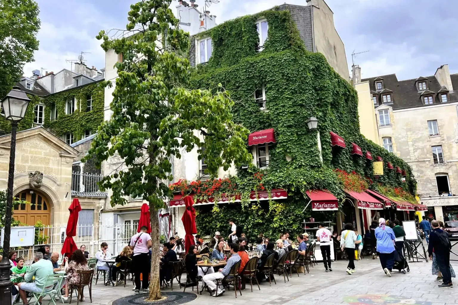 Building covered in green ivy and red awnings, with an outdoor restaurant patio in Paris. People are dining under a cloudy...