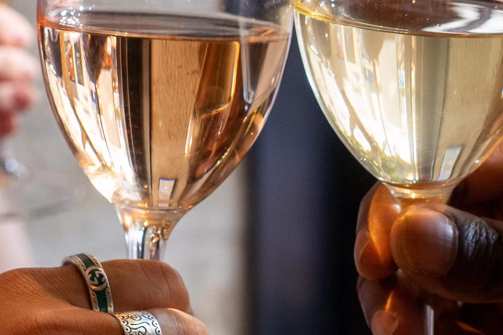 Two hands holding wine glasses, one with rosé wine and the other with white wine, clinking in a toast. One hand wears rings.