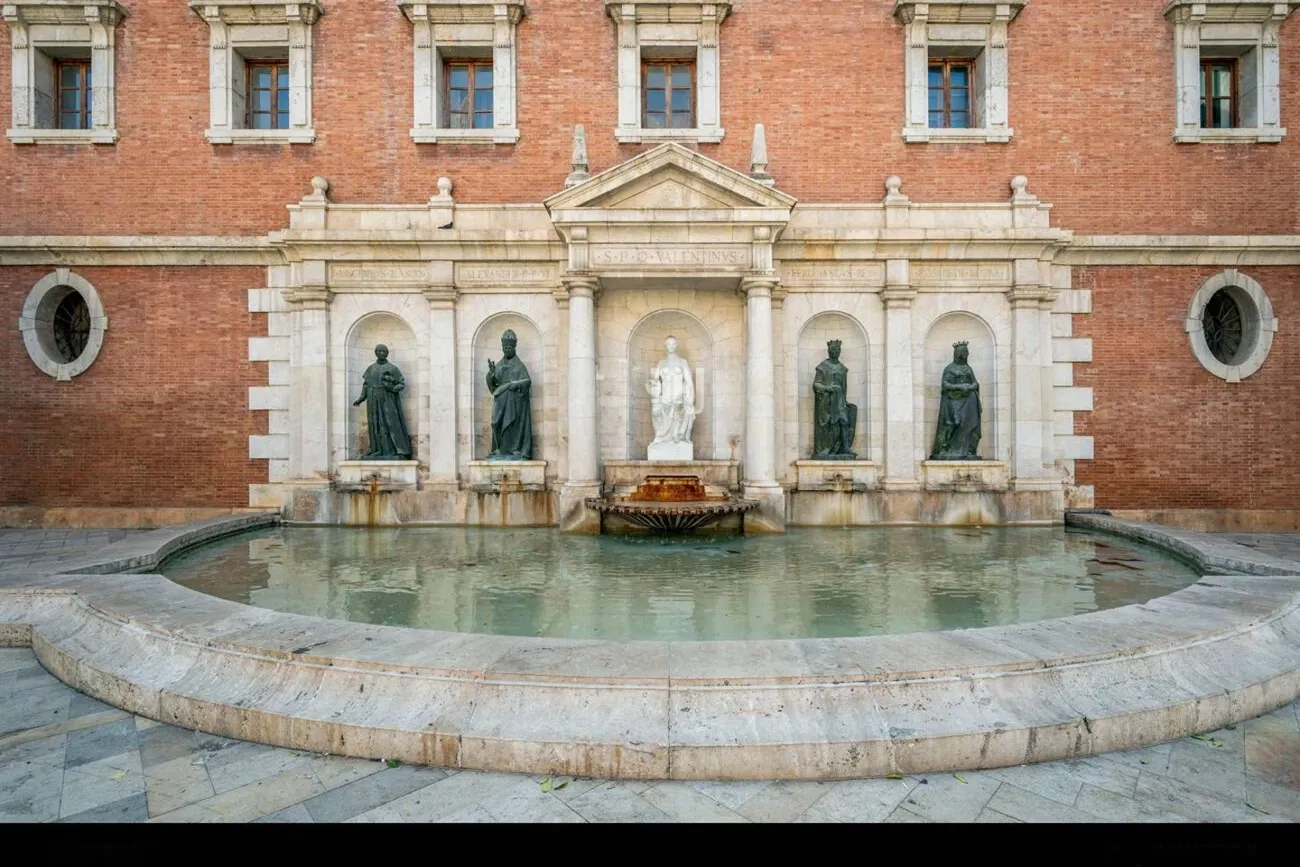 A wide, curved stone fountain in front of a brick building with statues in arched niches.