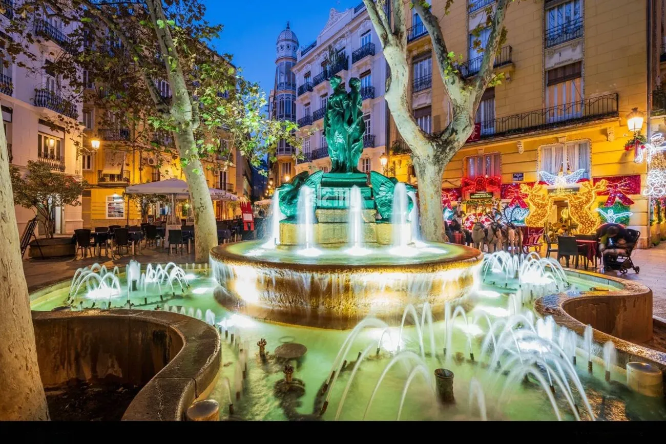 An illuminated fountain in Plaza Rodrigo Botet in Valencia, surrounded by trees and buildings with Christmas lights.