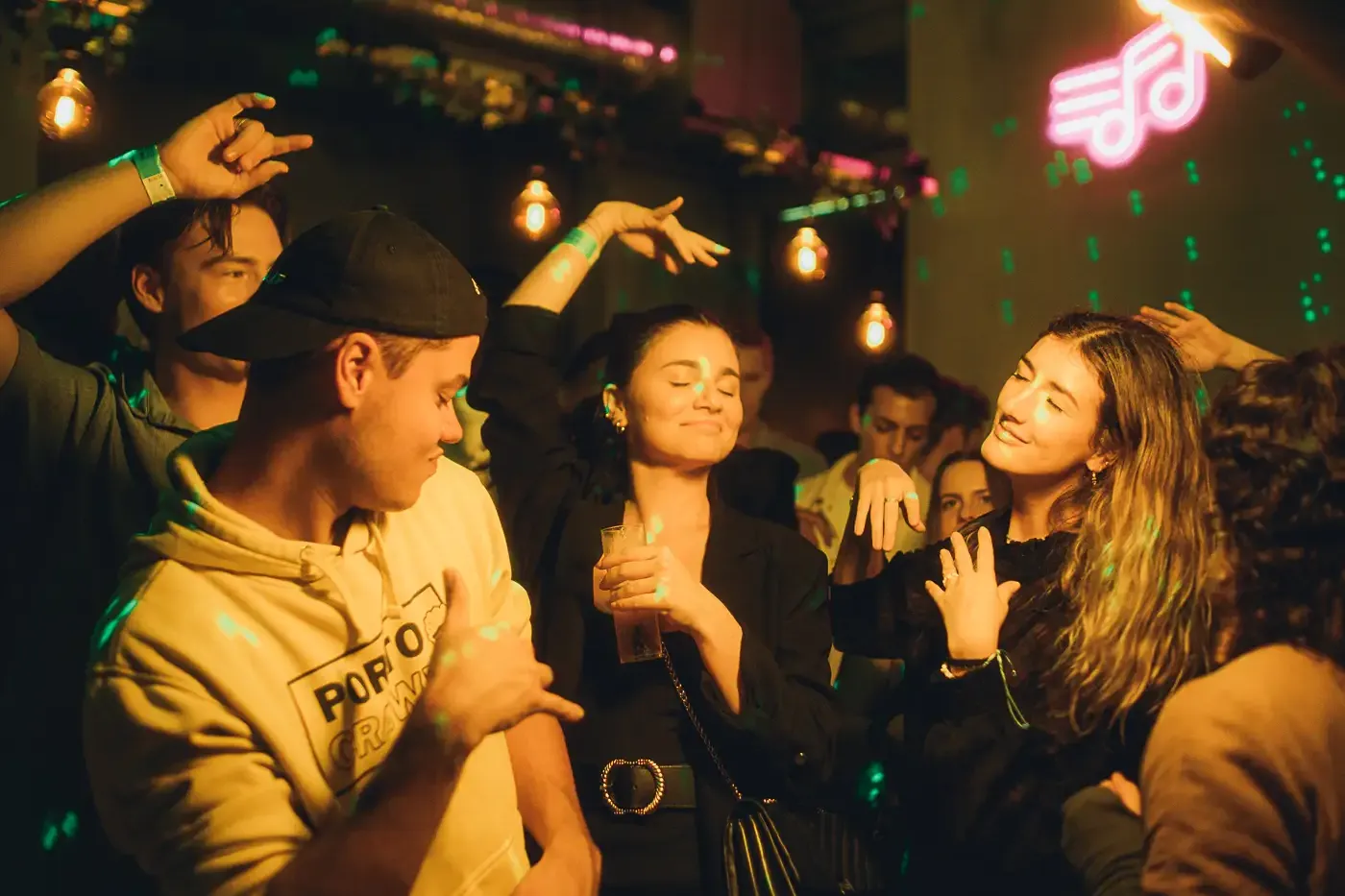 People dancing in a dimly lit club or bar, with green laser lights and a pink neon musical note sign.