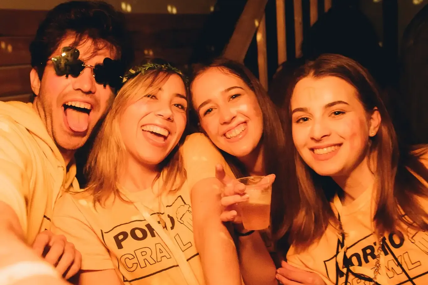 Four young people, two men and two women, smiling in a dimly lit bar. They wear yellow t-shirts with 'PORTO PUB CRAWL' and...