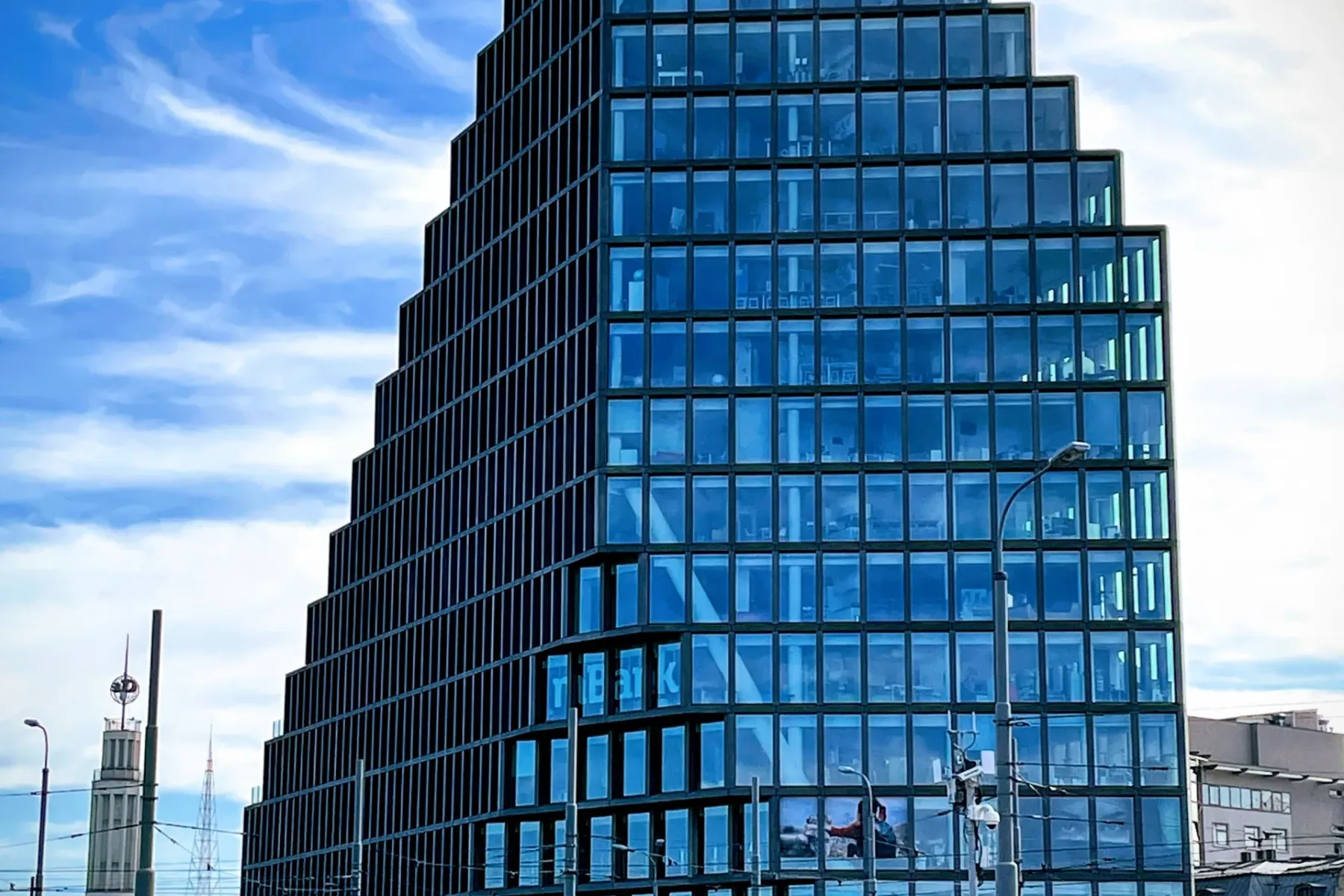 Modern Bałtyk building in Poznań with a stepped glass facade, set against a blue sky, with a street and historical tower v...