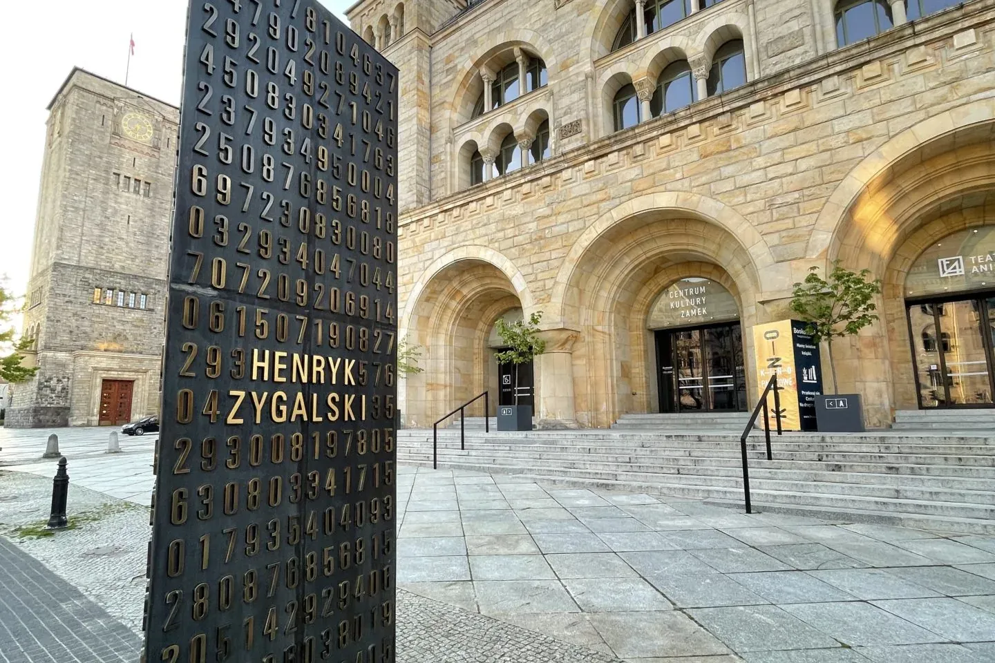 Dark monument with gold numbers and 'HENRYK ZYGALSKI' on a paved plaza, facing Poznan Imperial Castle with arches.