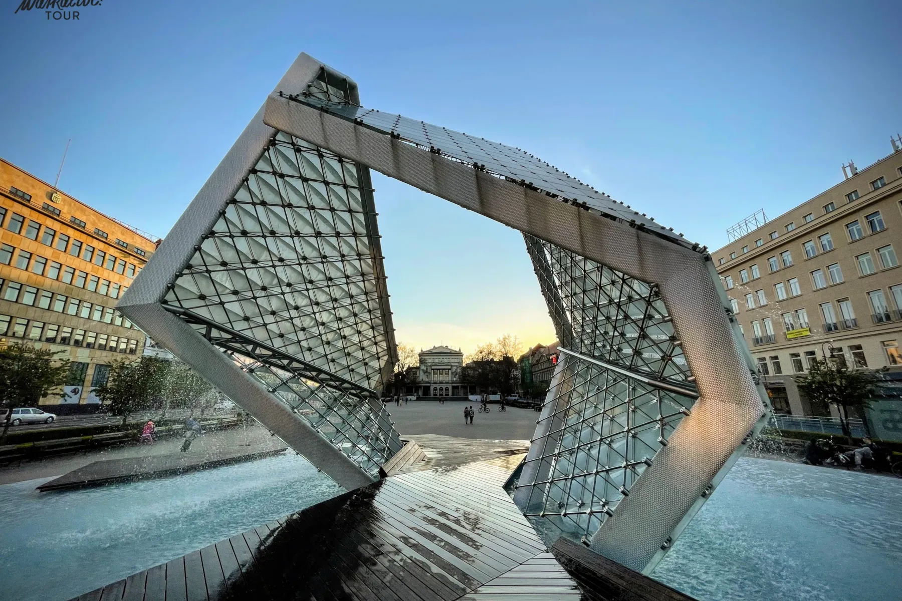 Modern glass and metal fountain with water jets, shaped like an open cube, in a city square at dusk. Buildings in the back...