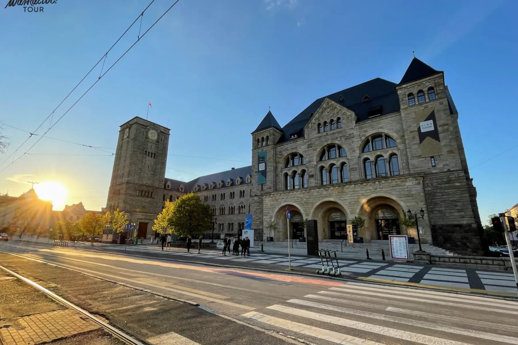 Stone Imperial Castle building in Poznań with a clock tower, tram tracks, and a crosswalk in the foreground. Sun is low on...
