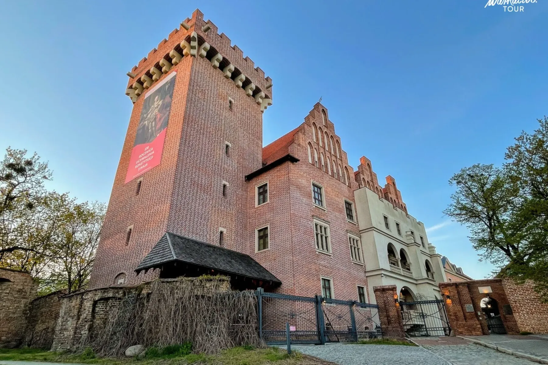 View of the Royal Castle in Poznań, a large brick building with a tall tower, gabled facade, and old wall, under a blue sky.