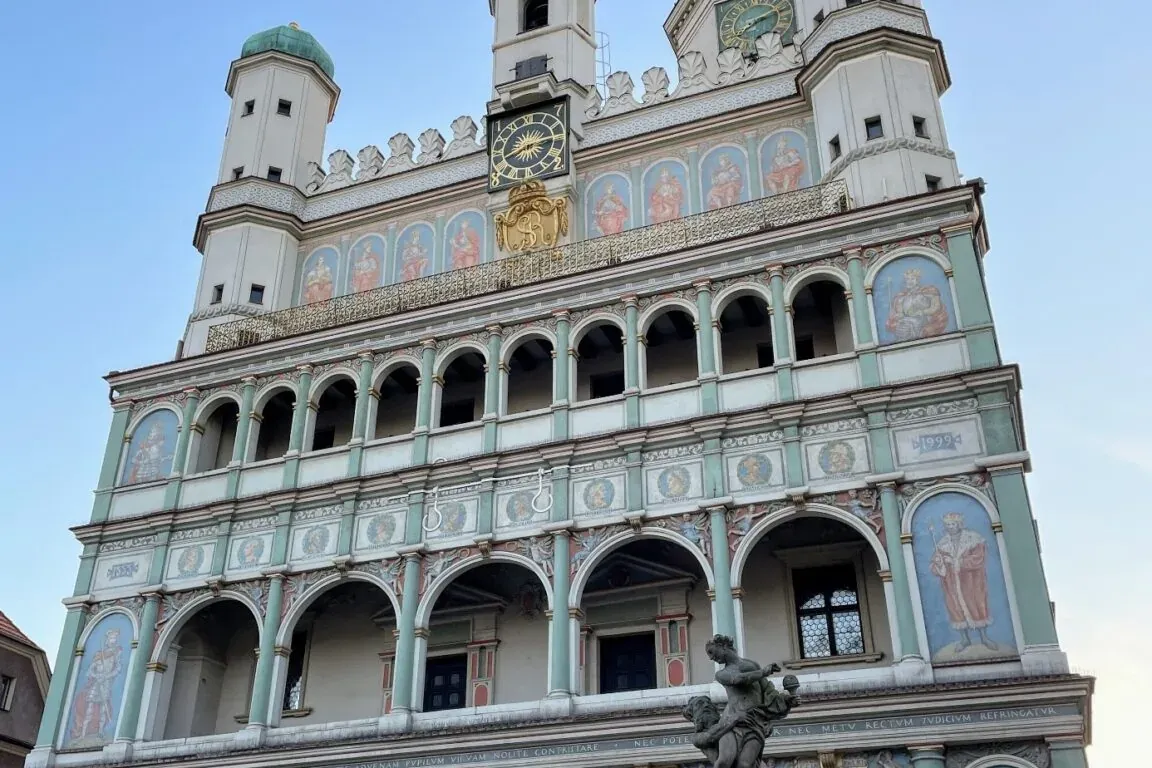 Renaissance Town Hall in Poznan with ornate facade, arcades, frescoes, clocks, and green spires under a blue sky, fountain...