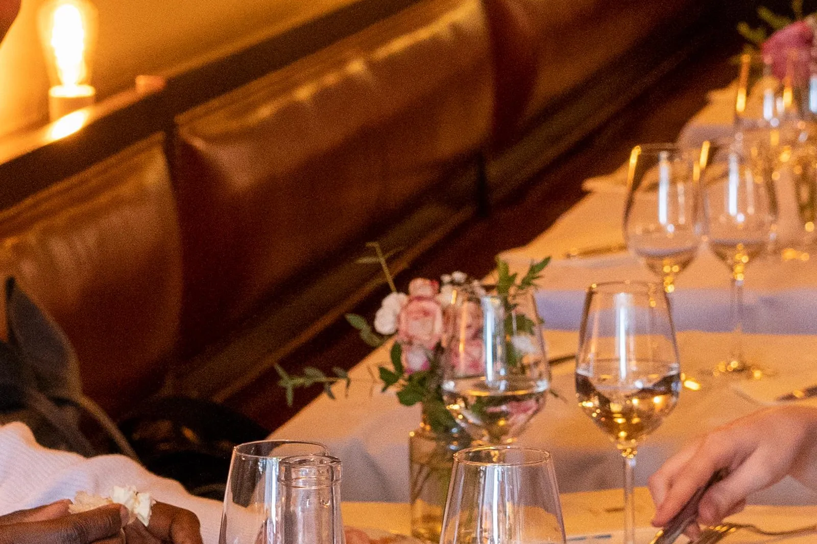 People dining at a restaurant table with bread, a water bottle, wine glasses, and a cheese board. Warm interior lighting.