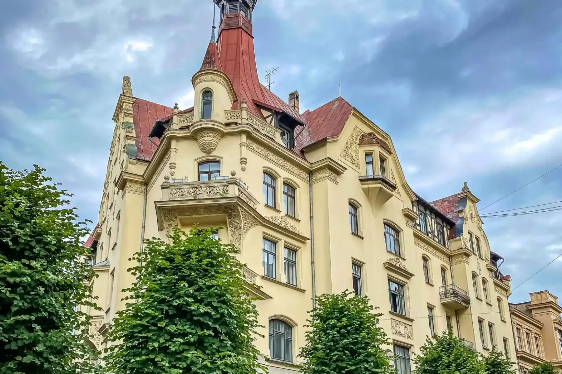 Ornate Art Nouveau building in Riga with a yellow facade, red roof, and central spire, framed by green trees against a clo...