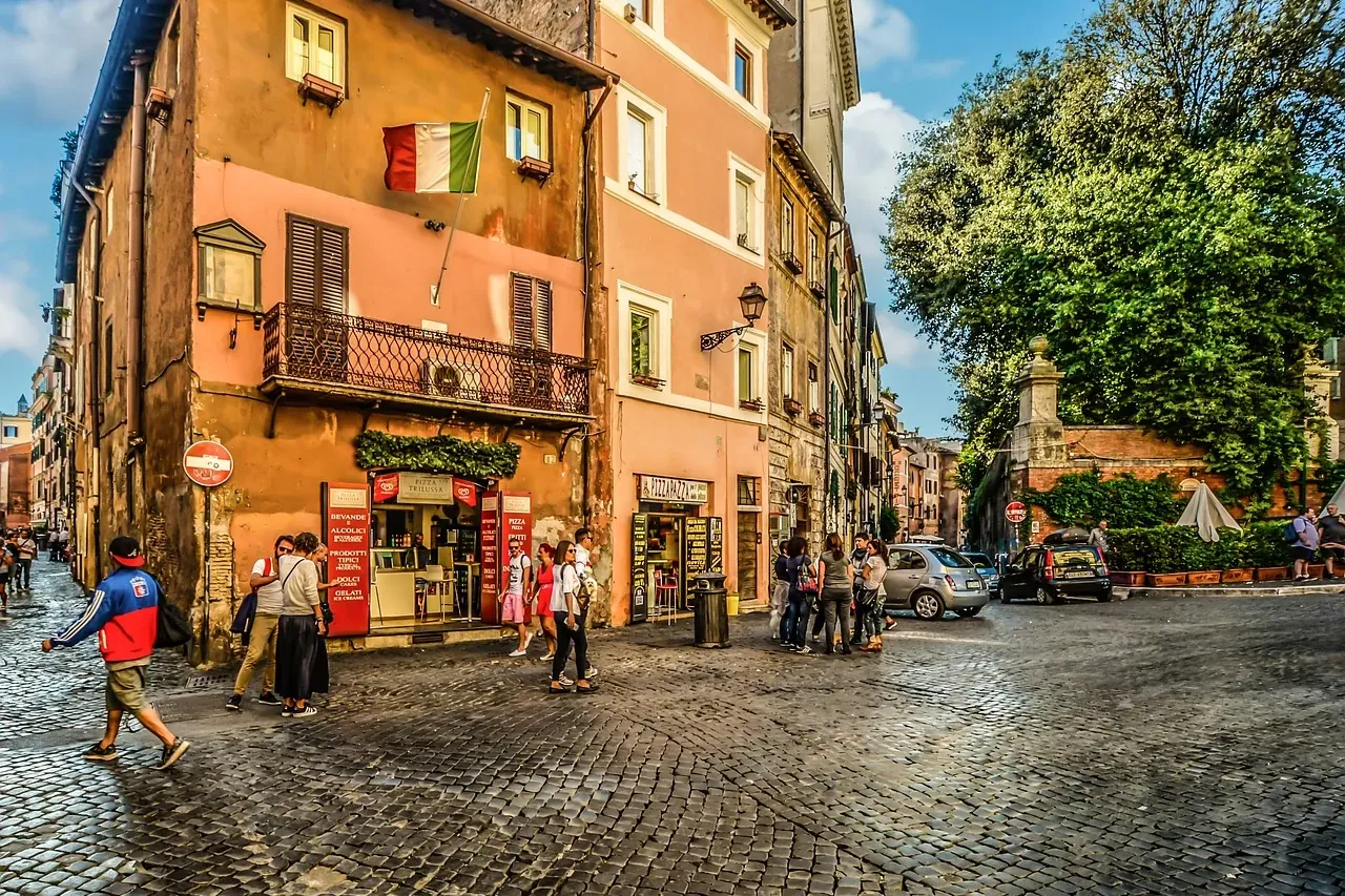 Cobblestone street in Rome with historic buildings, an Italian flag, people walking, and parked cars under a blue sky.