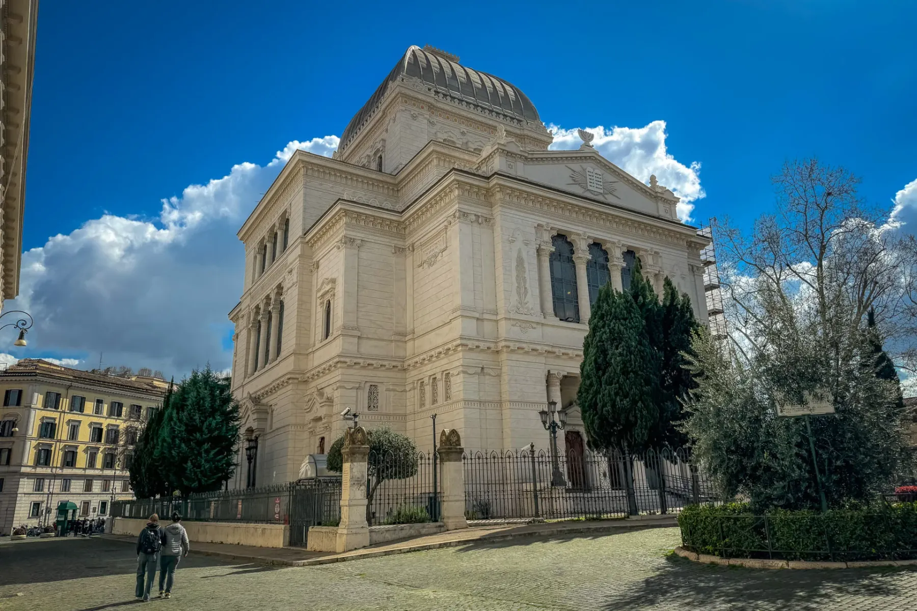 The Great Synagogue of Rome with its dome, a cobblestone street, green trees, and two people walking under a bright blue sky.