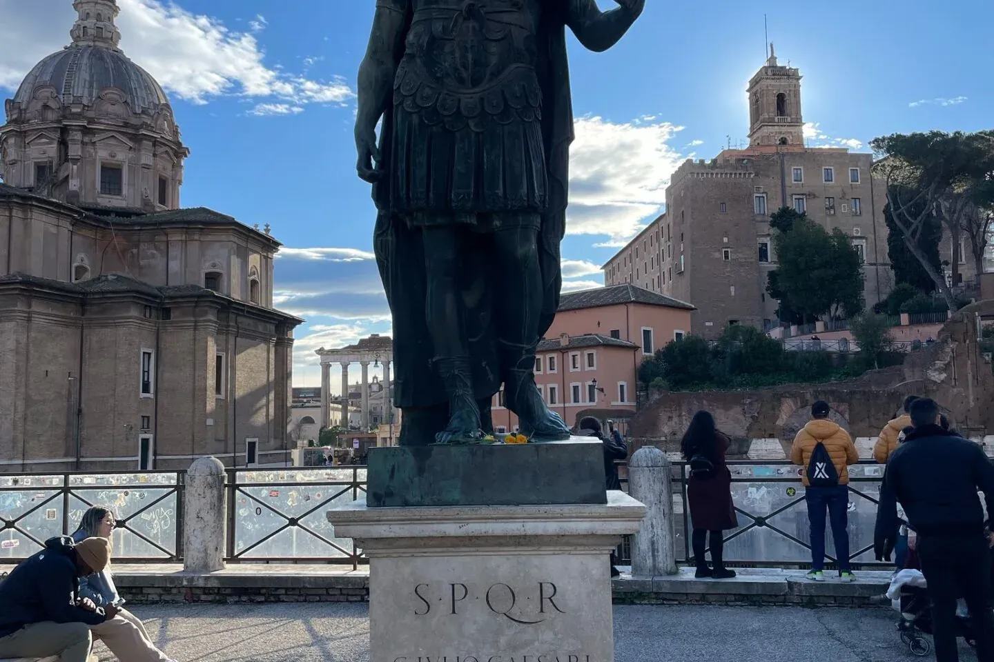 Bronze statue of Julius Caesar in Rome, with Roman Forum ruins and a domed church under a blue sky.