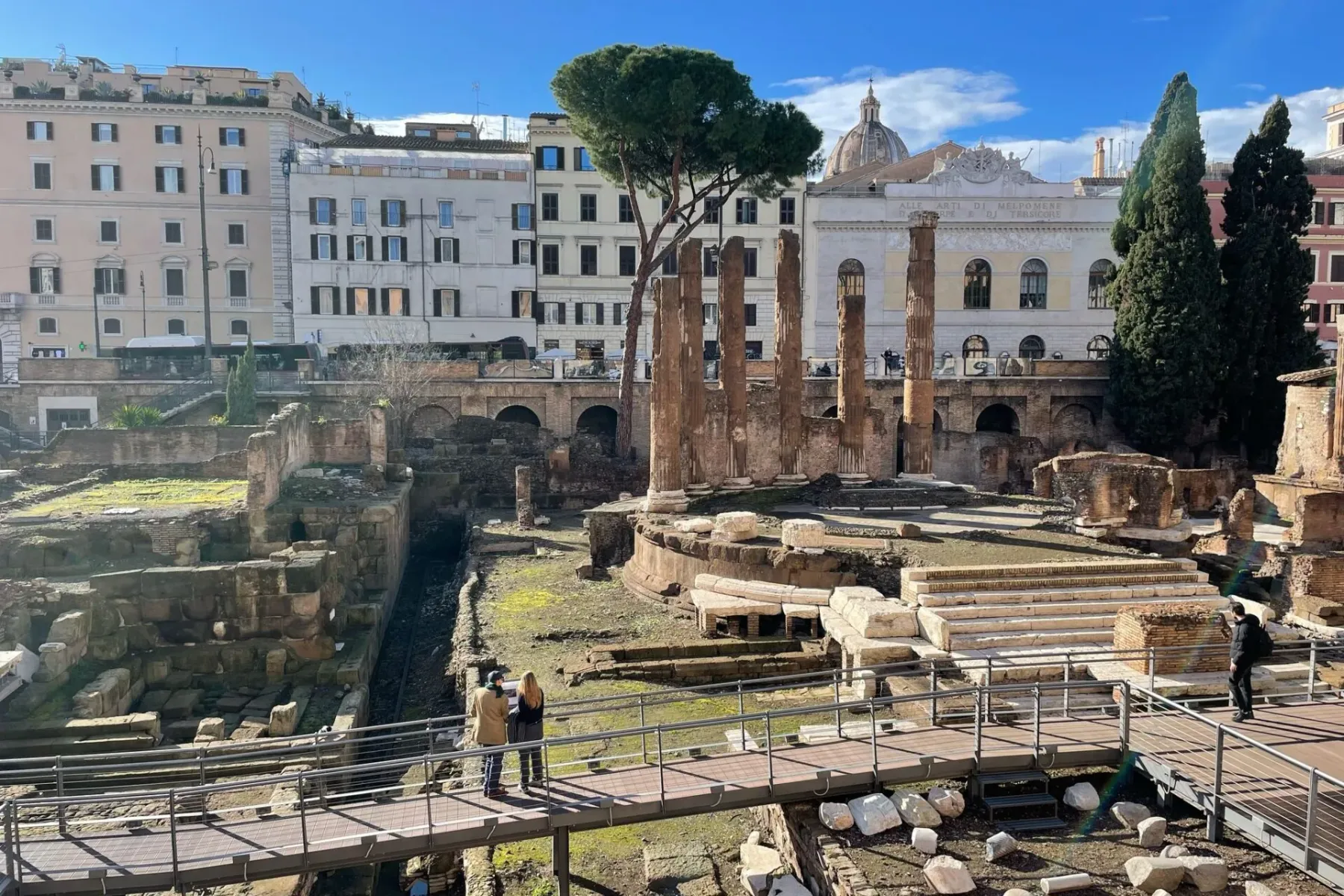 View of ancient Roman ruins with excavated levels, stone columns, and a modern wooden walkway. City buildings in background.