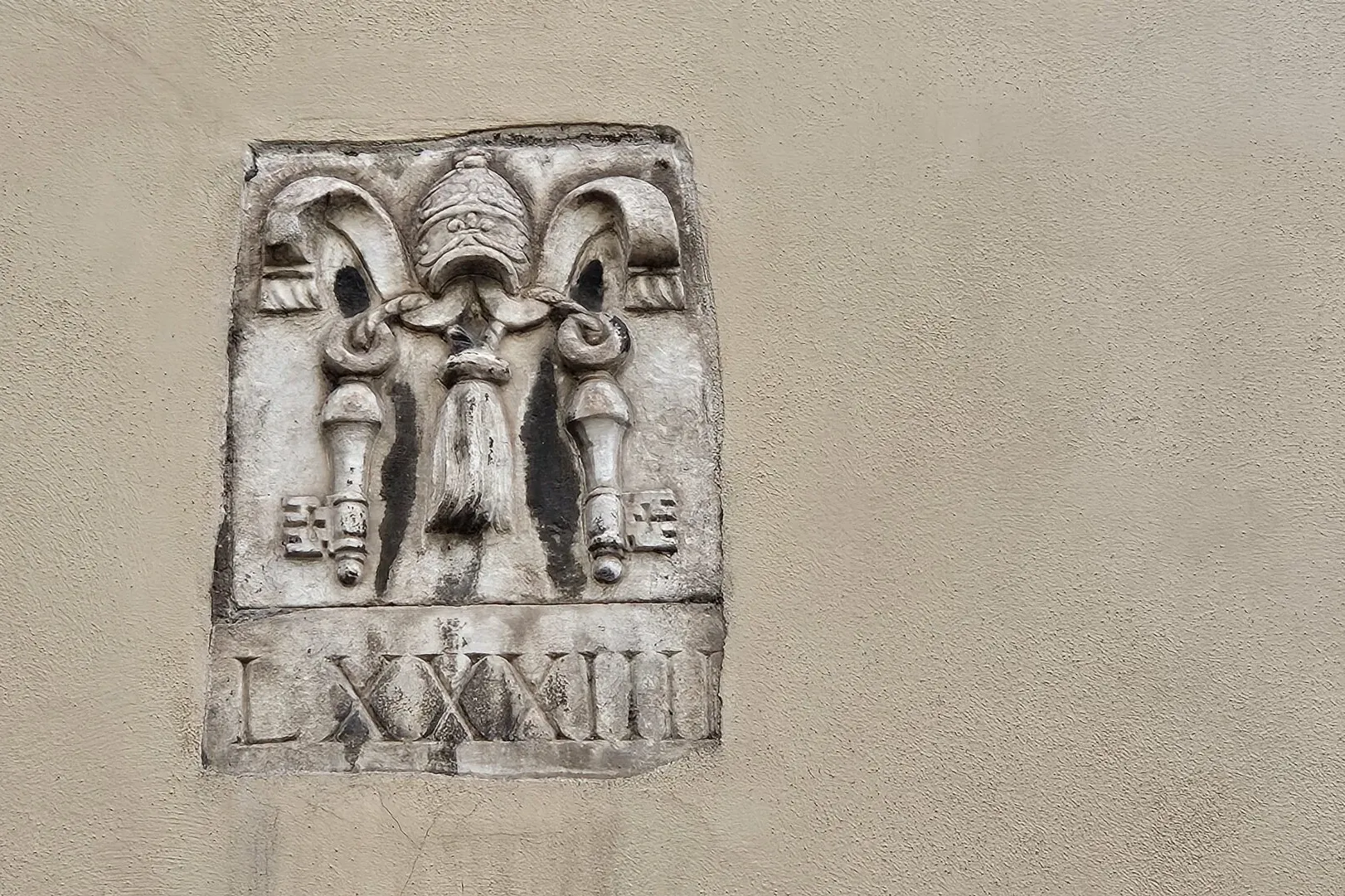 Stone relief on a textured wall showing a papal tiara, crossed keys, and tassels, with Roman numerals LXXXIII below.