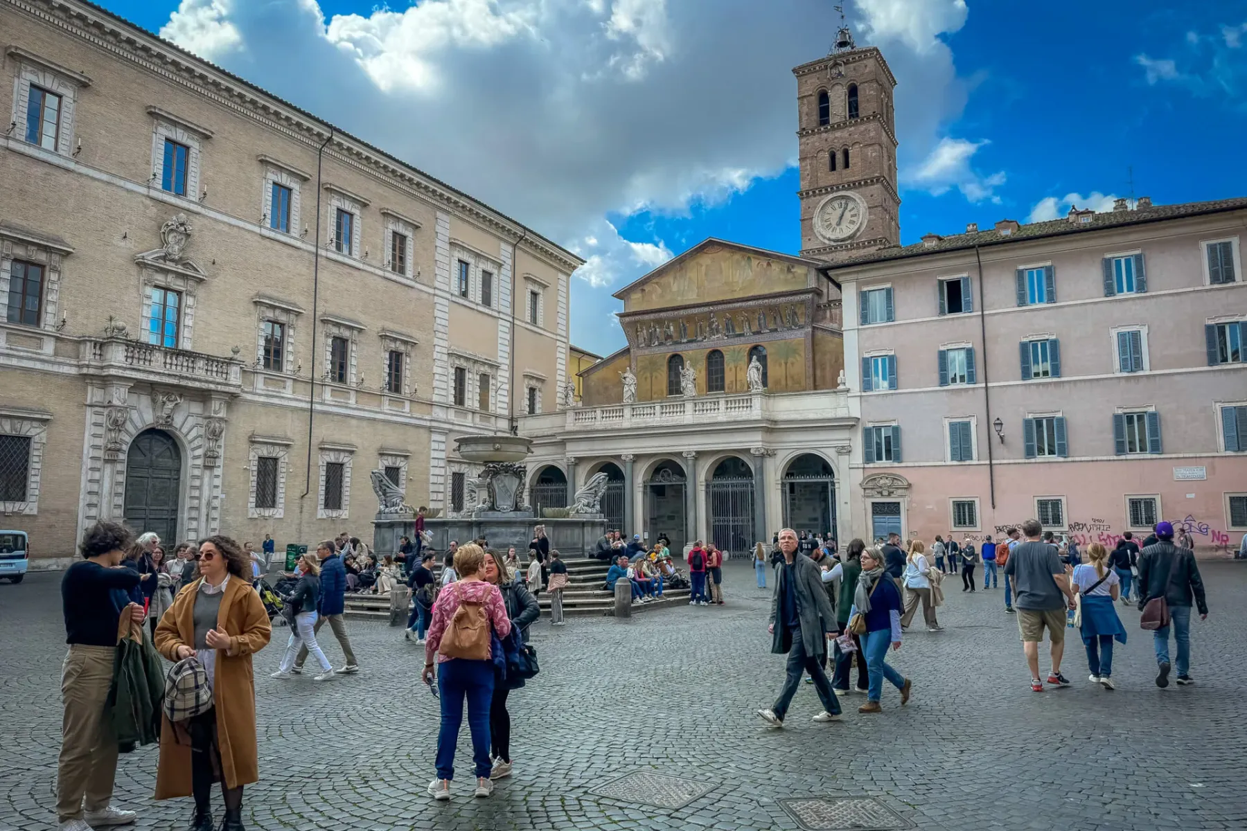 Cobblestone Piazza Santa Maria in Trastevere, Rome, with a basilica, bell tower, fountain, and many people walking.