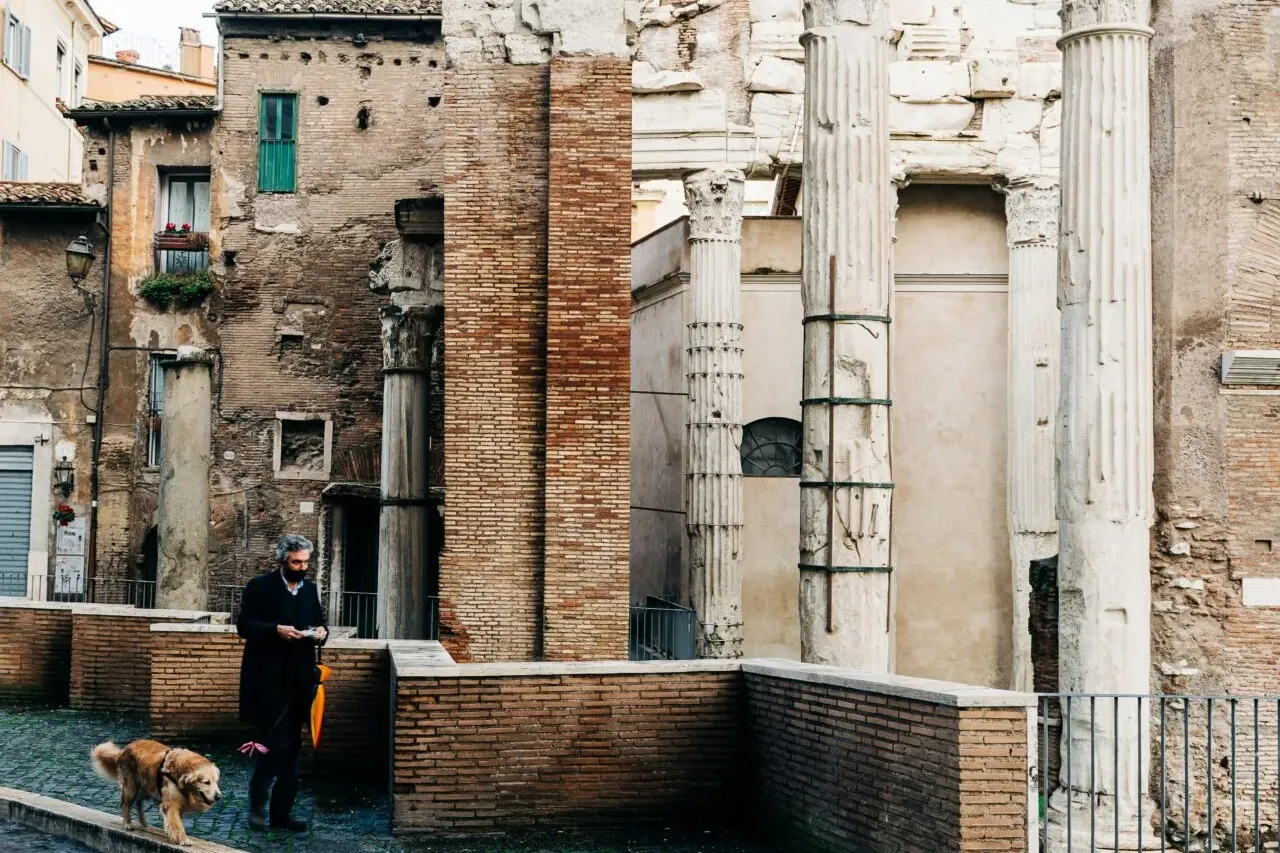 Man walking a golden retriever on a wet cobblestone street past ancient Roman ruins with tall columns and Latin inscriptions.