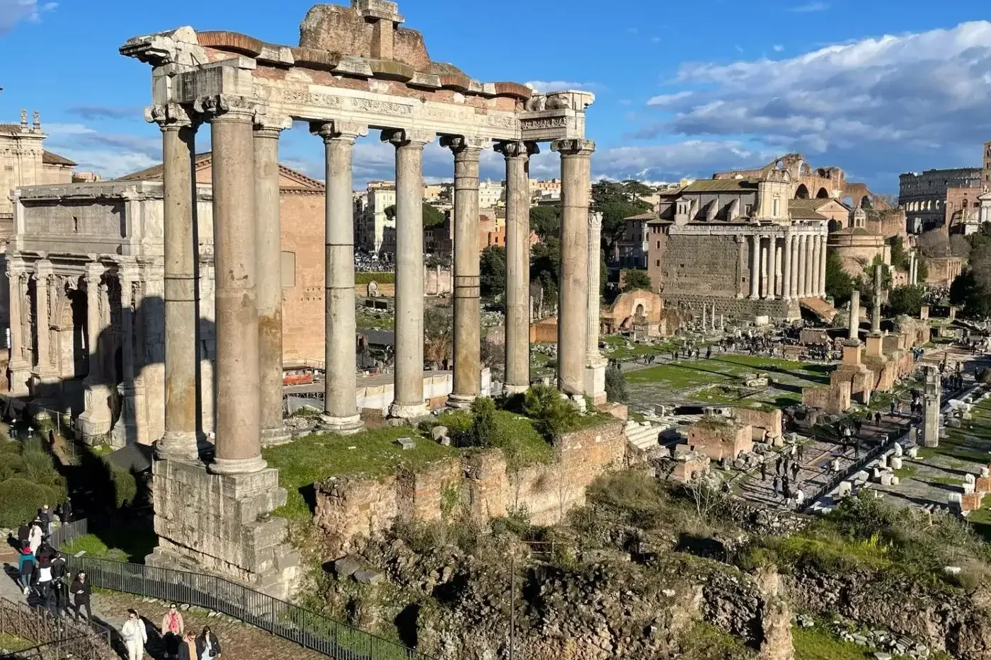 View of the Roman Forum in Rome, showing ancient ruins including eight tall columns of the Temple of Saturn under a blue sky.