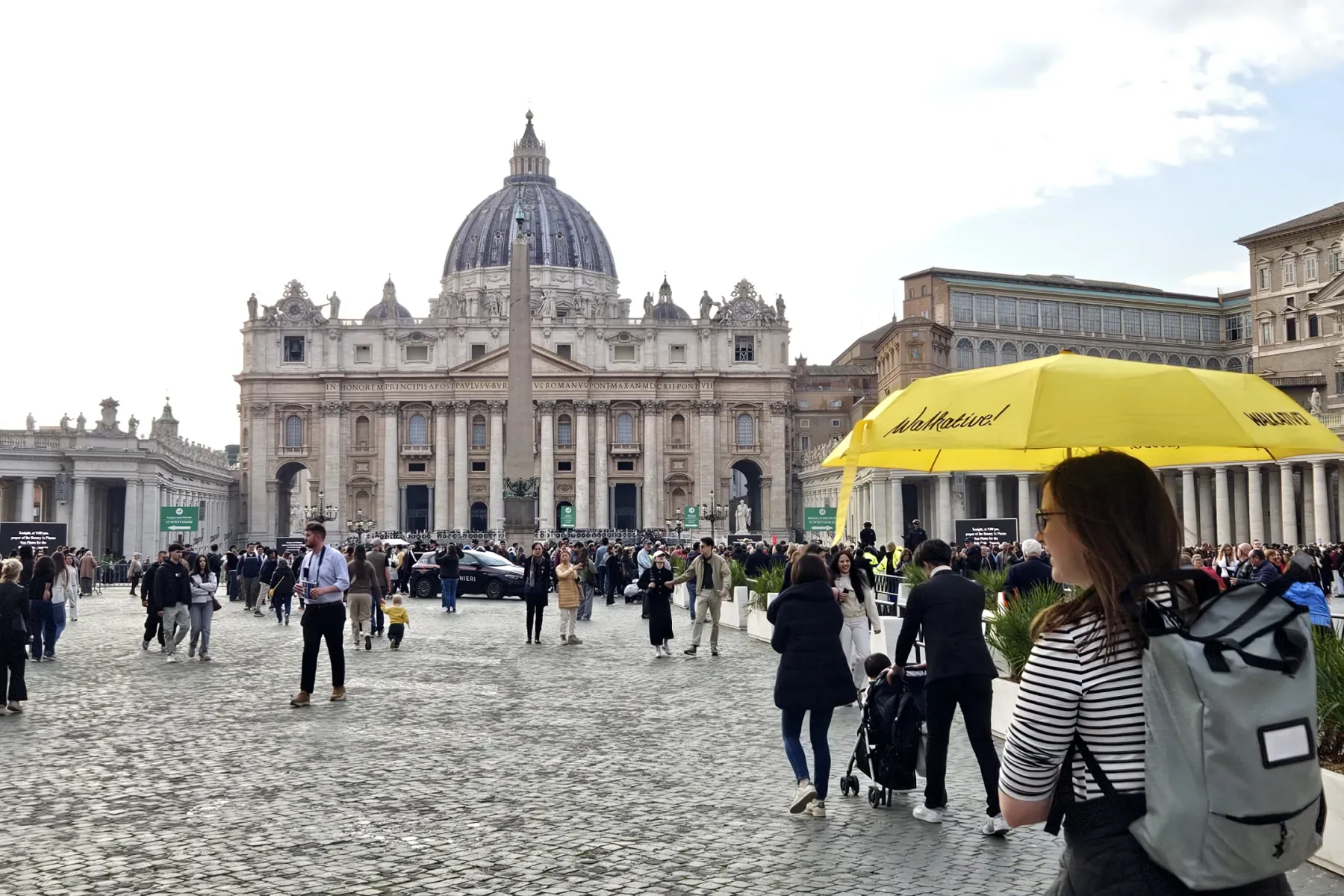Crowded St. Peter's Square with St. Peter's Basilica, an obelisk, and colonnades under a partly cloudy sky. A yellow Walka...