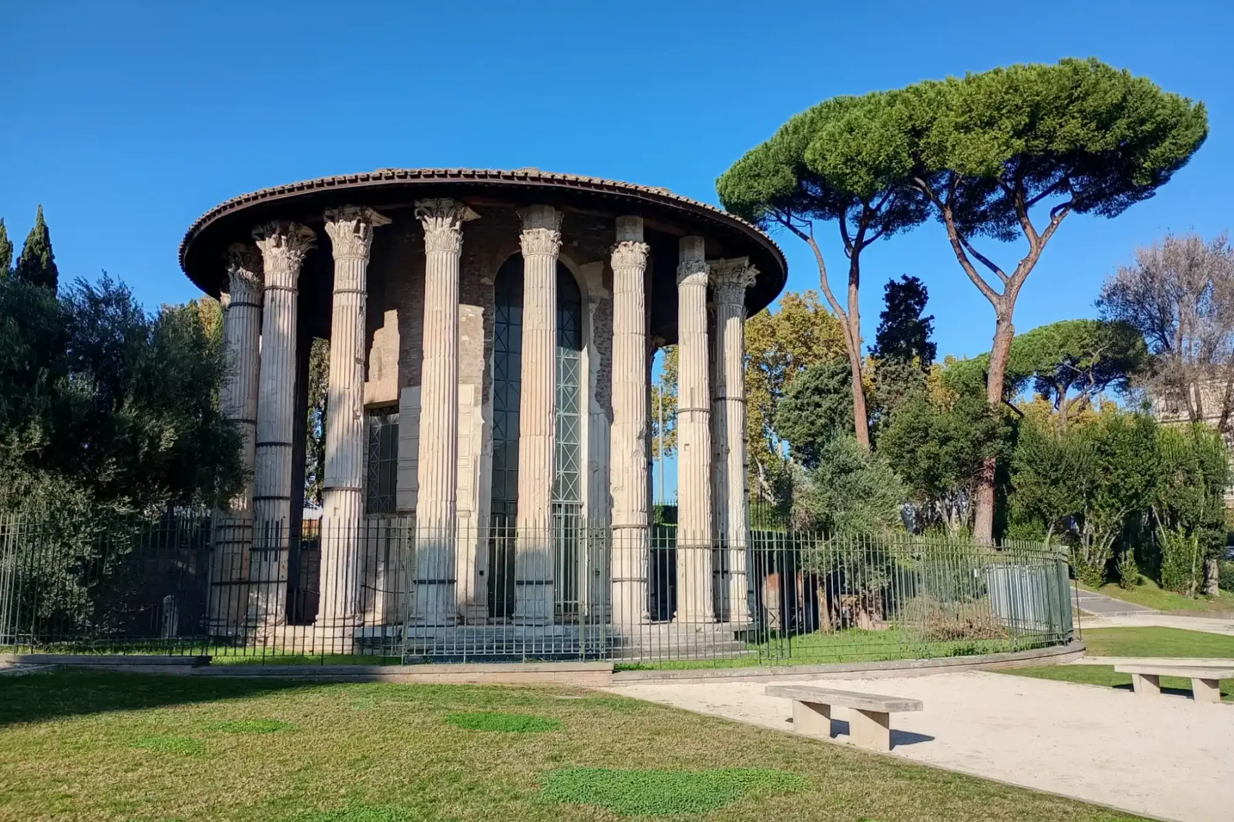 Ancient round Temple of Hercules Victor in Rome featuring Corinthian columns, surrounded by trees and a metal fence.
