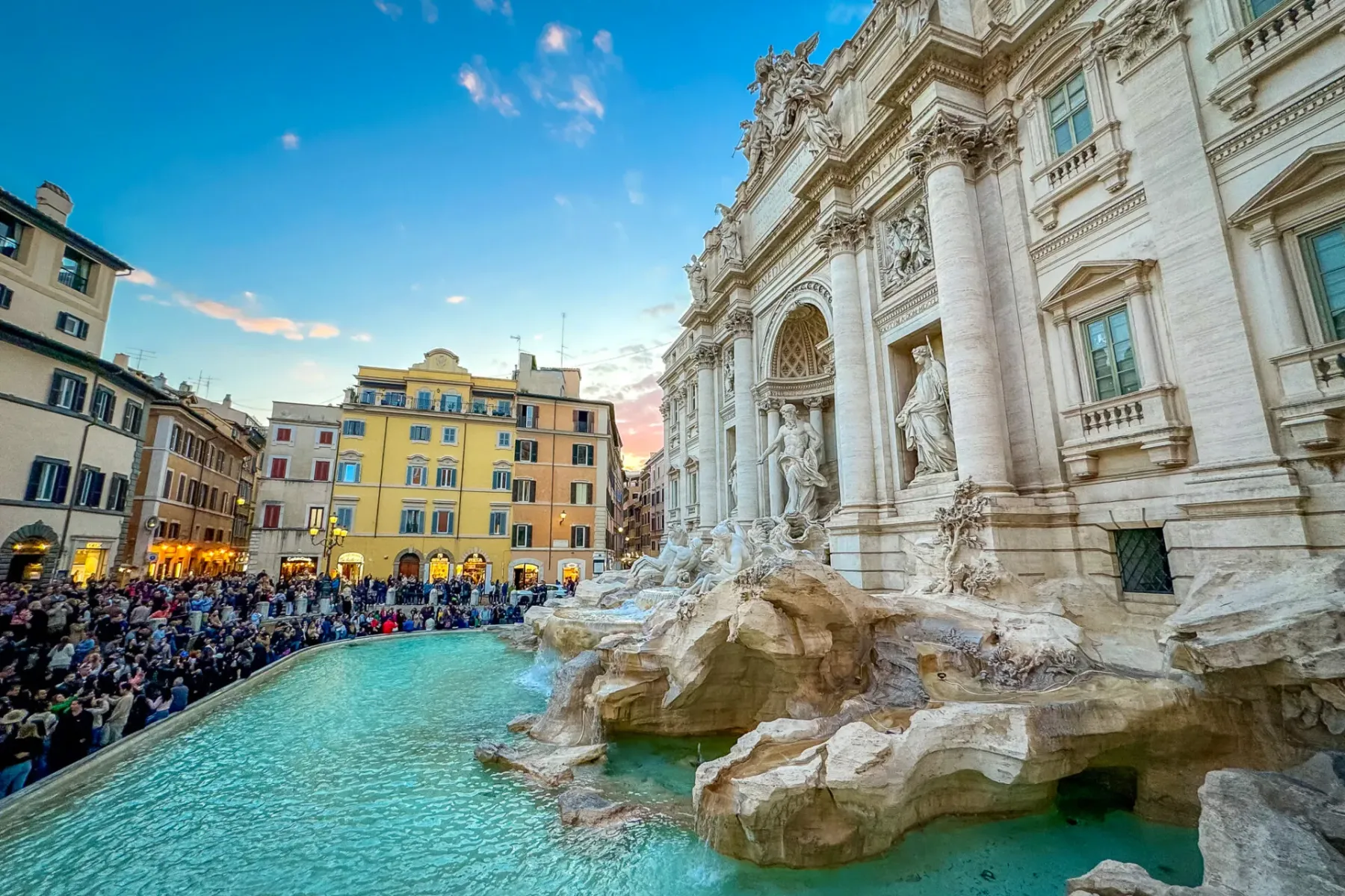 Crowded Trevi Fountain in Rome at dusk, with baroque sculptures, turquoise water, and historical buildings under a pink-bl...