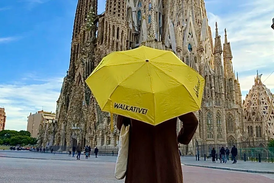 Person holding a yellow Walkative umbrella, facing the intricate spires of Sagrada Família under a blue sky in Barcelona.