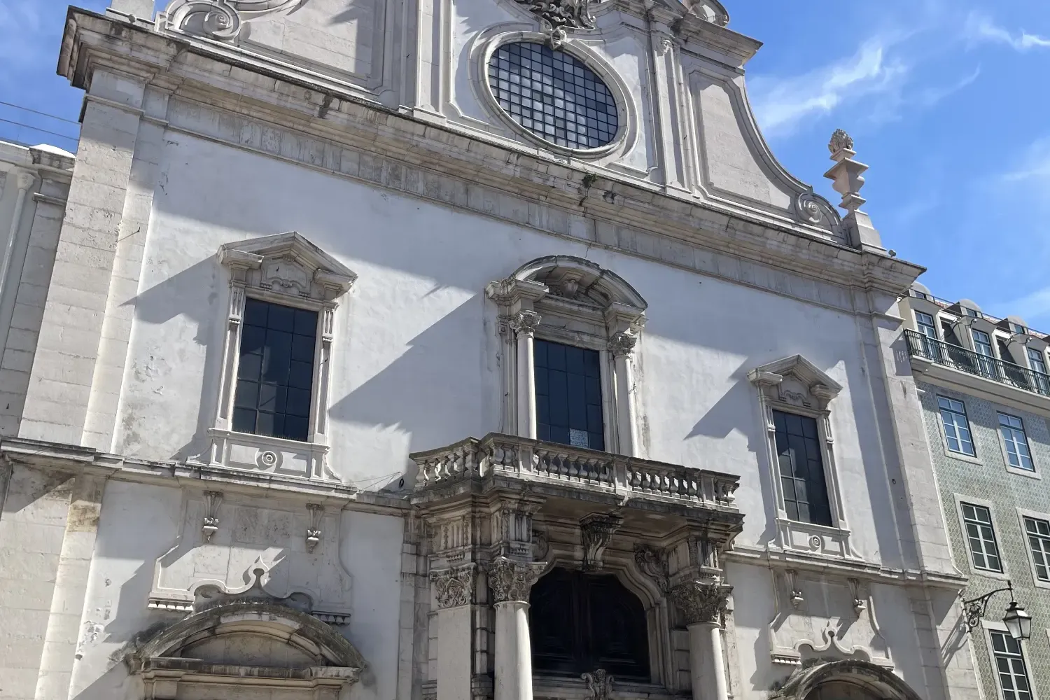 Ornate white facade of São Domingos Church in Lisbon, with large dark doors, a balcony, and a cross on the roof. People ga...