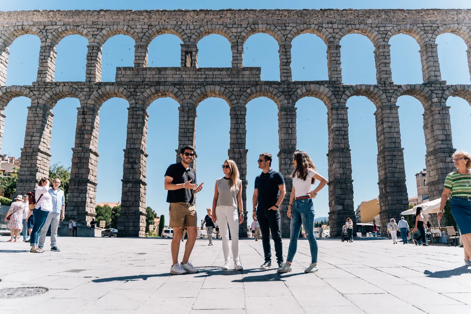 A tour guide talks to three tourists in front of the large, sunny Aqueduct of Segovia in Spain.