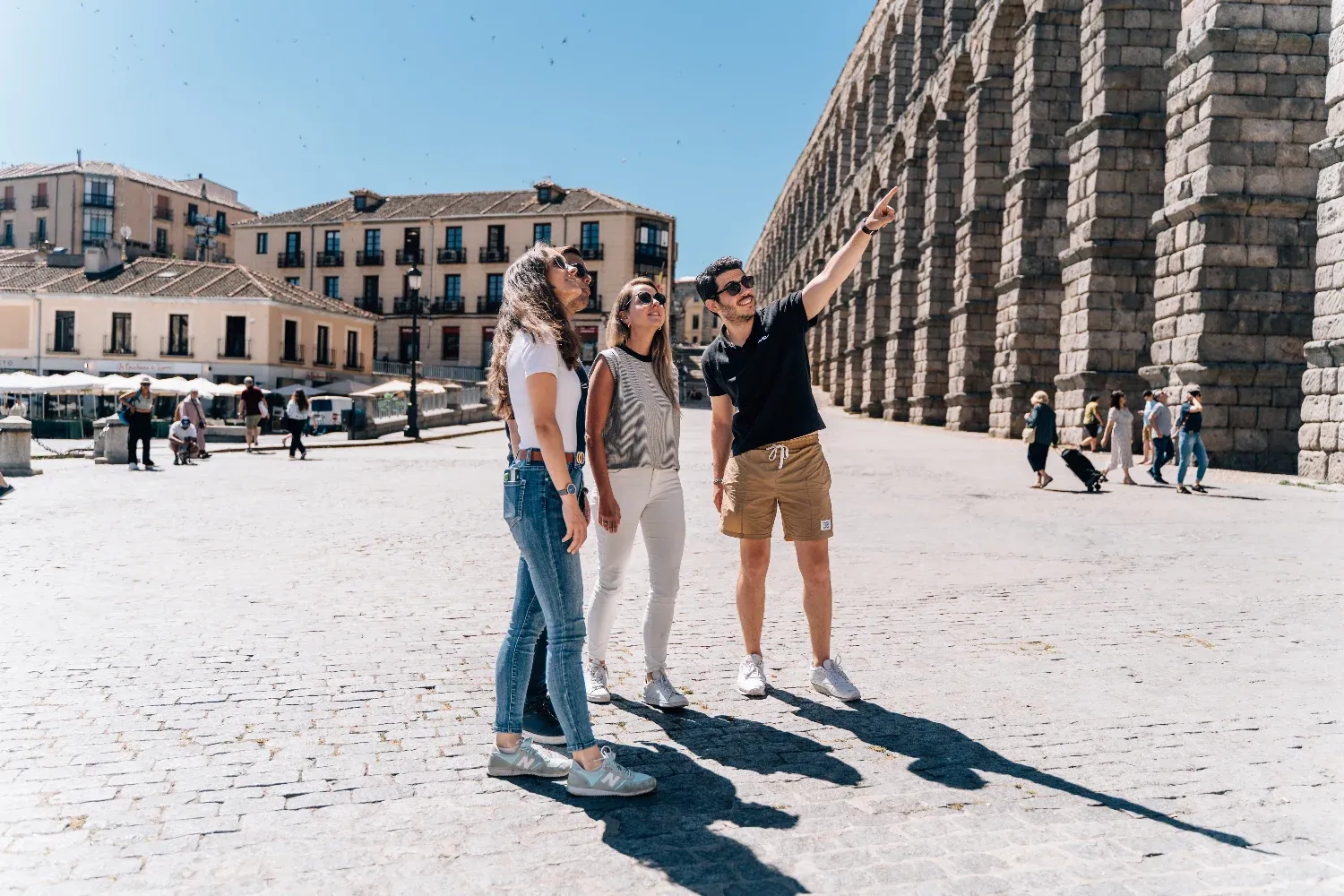 A tour guide points up at the Aqueduct of Segovia for three tourists on a sunny day.
