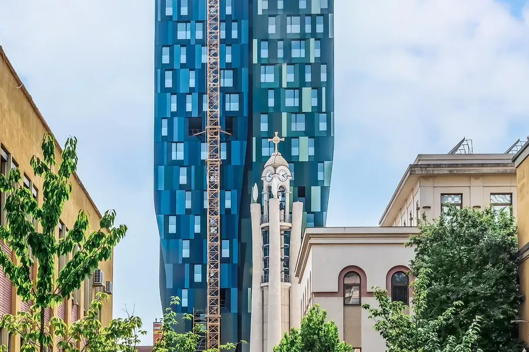 Modern blue skyscraper with a yellow construction crane, behind a traditional church bell tower and older buildings in Tir...