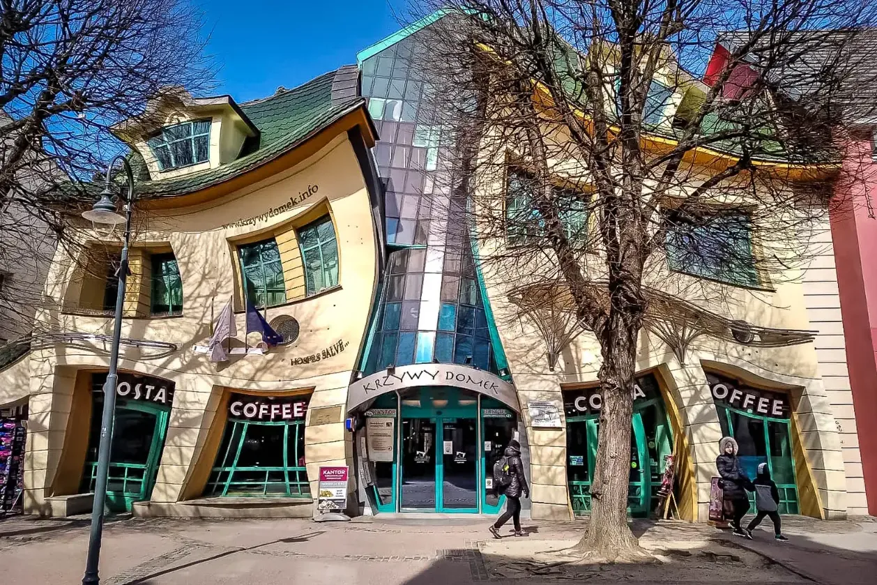 The Crooked House (Krzywy Domek) in Sopot, with a wavy facade and green roof, shops on the ground floor, and bare trees.
