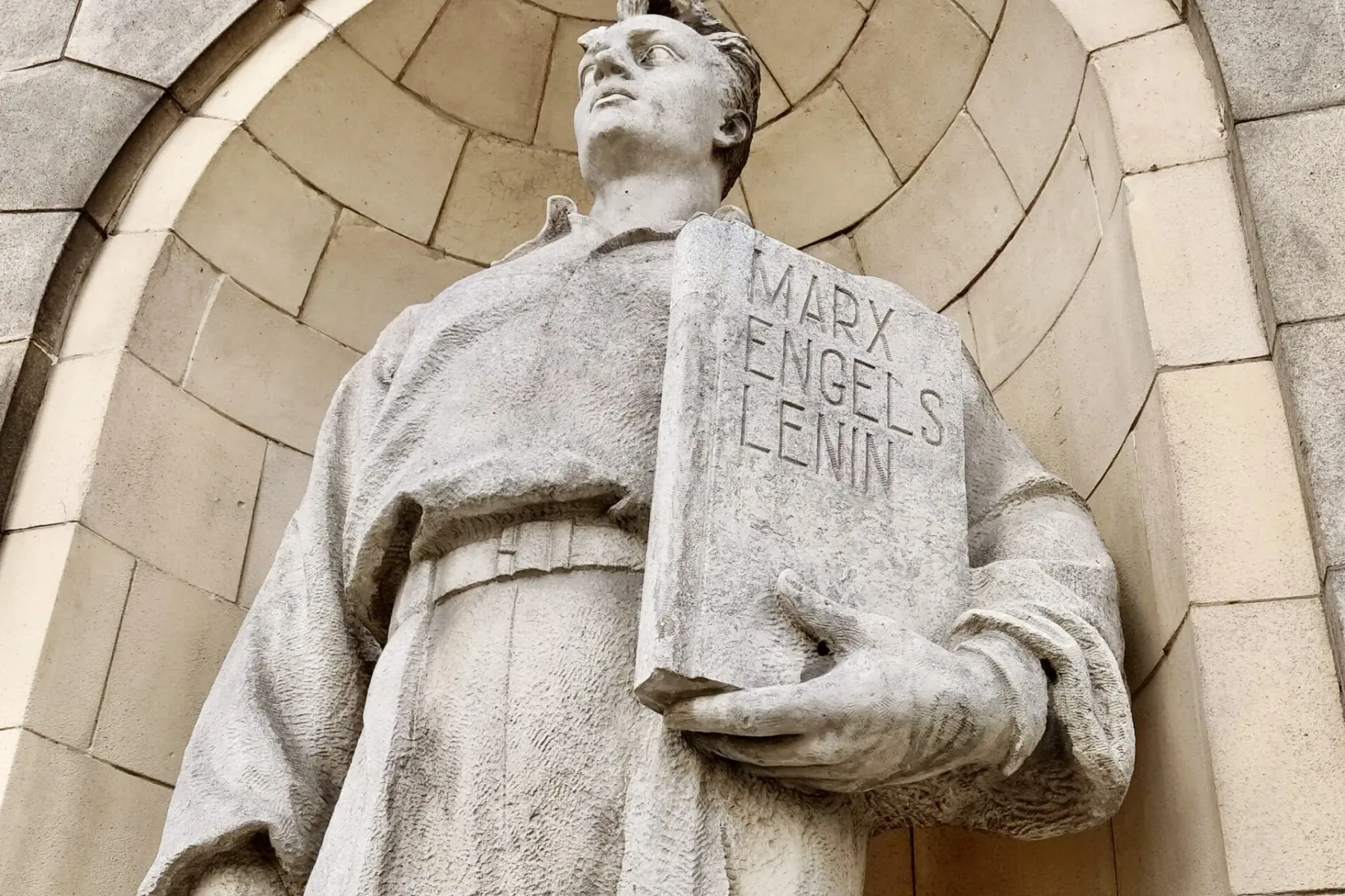 Stone statue of a man looking up, holding a book with "MARX ENGELS LENIN" carved on its cover, under an arched stone recess.