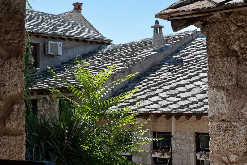 View from a shaded balcony with a wooden railing, showing traditional stone-tiled roofs, a cobblestone courtyard, and gree...